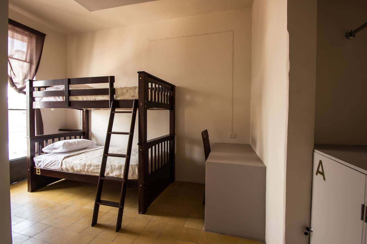 Cama litera con escalera, un escritorio y un armario en una habitación con paredes blancas y ventana con cortinas.