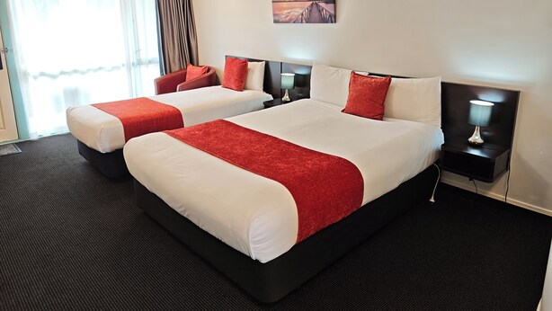 A hotel room with two beds, each with red and white bedding, a nightstand with a lamp, a painting on the wall, and a view of the outdoors through the window. - Great Lake Motel Taupo (Taupo)