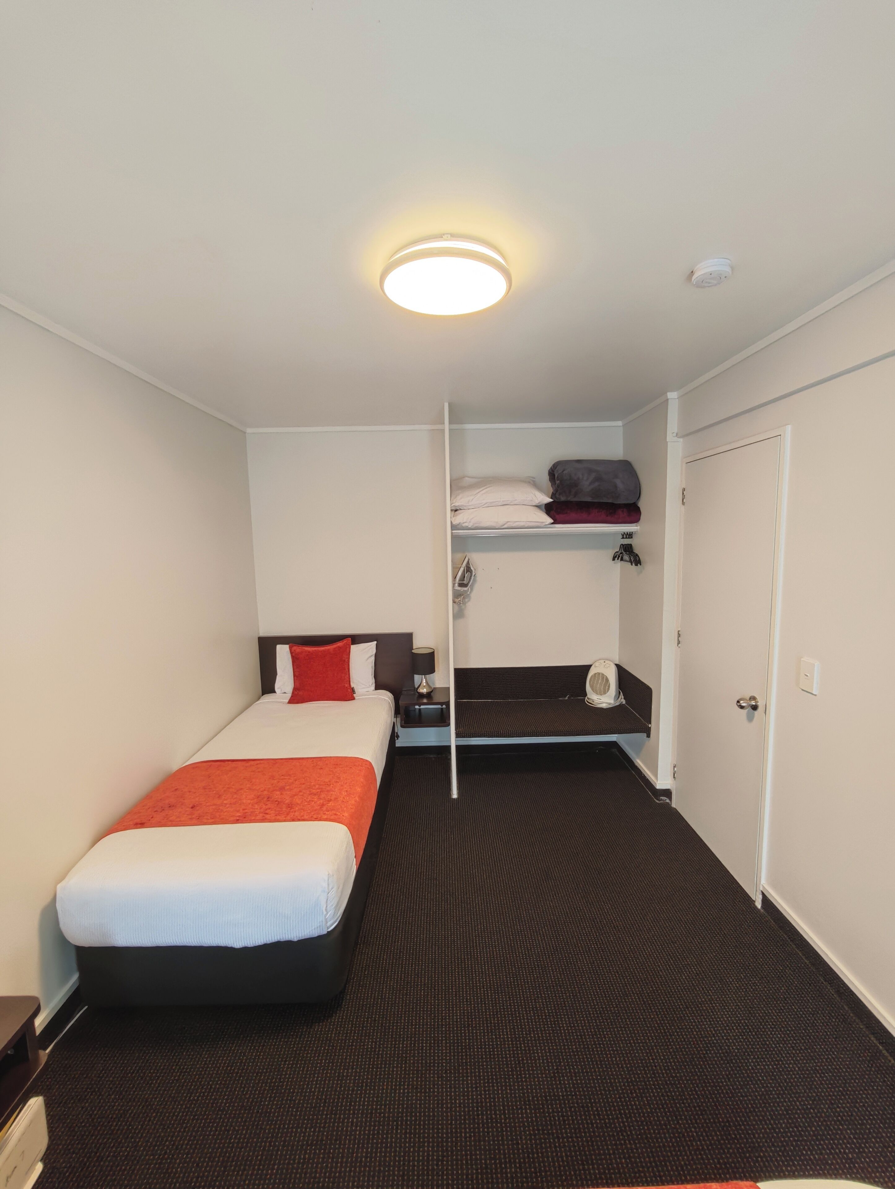 A hotel room with a bed, a small desk, a shelf with folded clothes, and a door.