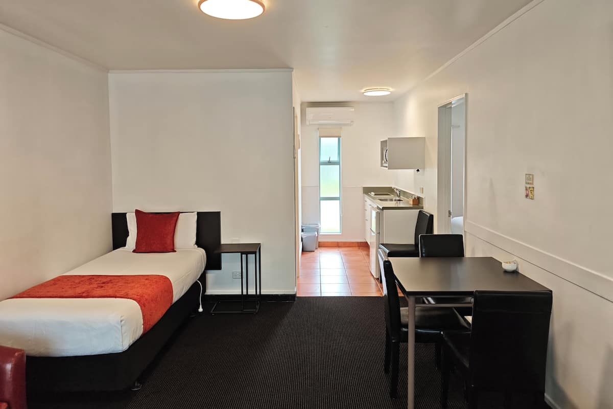 Une chambre d’hôtel de taille réduite, comprenant un lit simple, un bureau, une chaise, une petite table, un miroir et une kitchenette.
