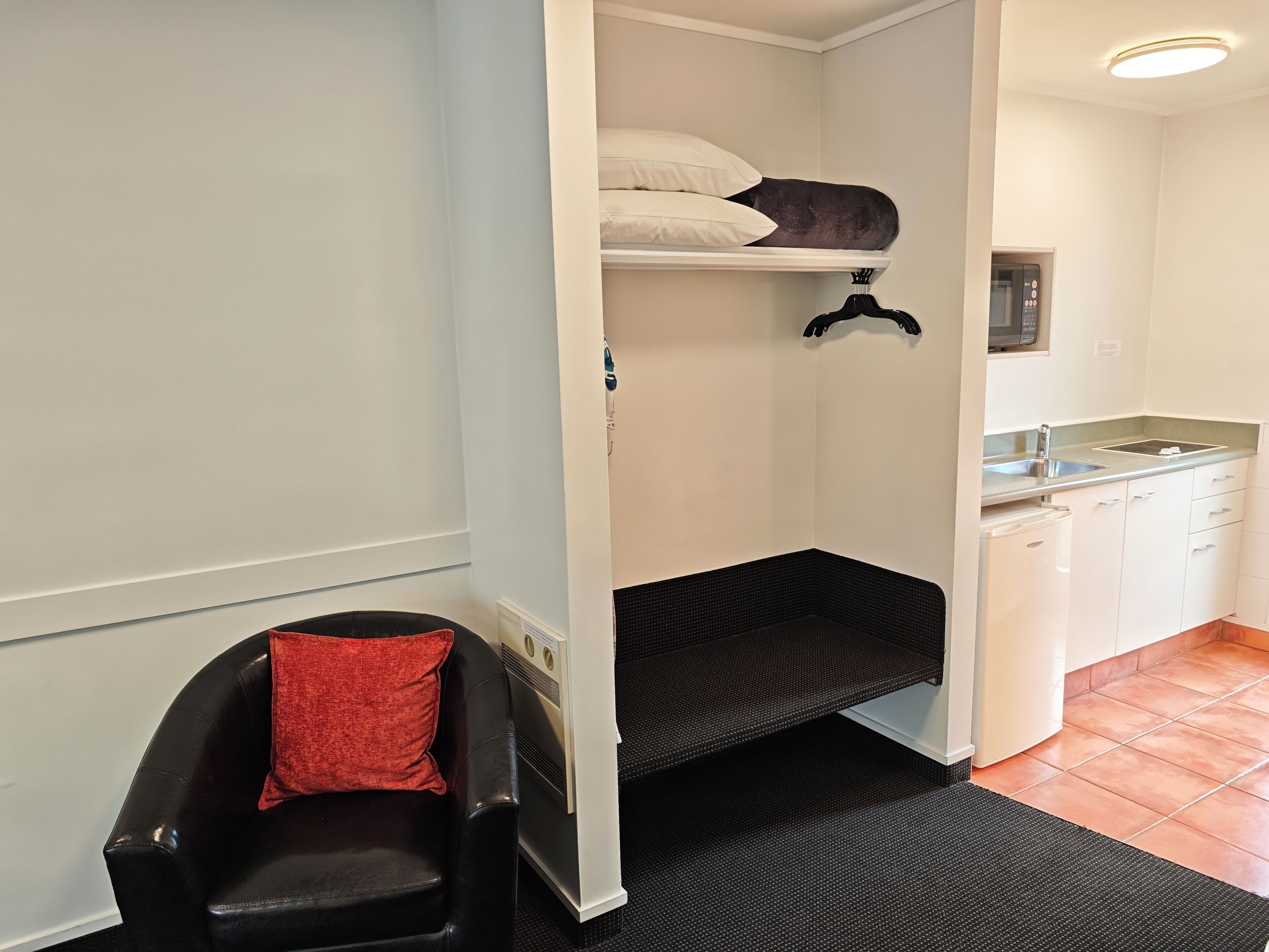 Habitación pequeña con una litera, un rincón de descanso con un cojín rojo, y una kitchenette con microondas y un lavabo.