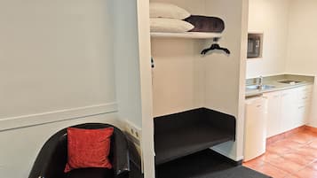Habitación pequeña con una litera, un rincón de descanso con un cojín rojo, y una kitchenette con microondas y un lavabo.