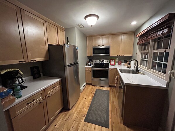 A compact kitchen with wooden cabinets, stainless steel appliances, and a window with blinds.