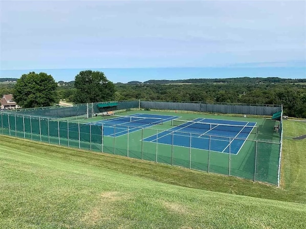 A tennis court complex with multiple courts, surrounded by a green fence, and set against a backdrop of trees and hills.