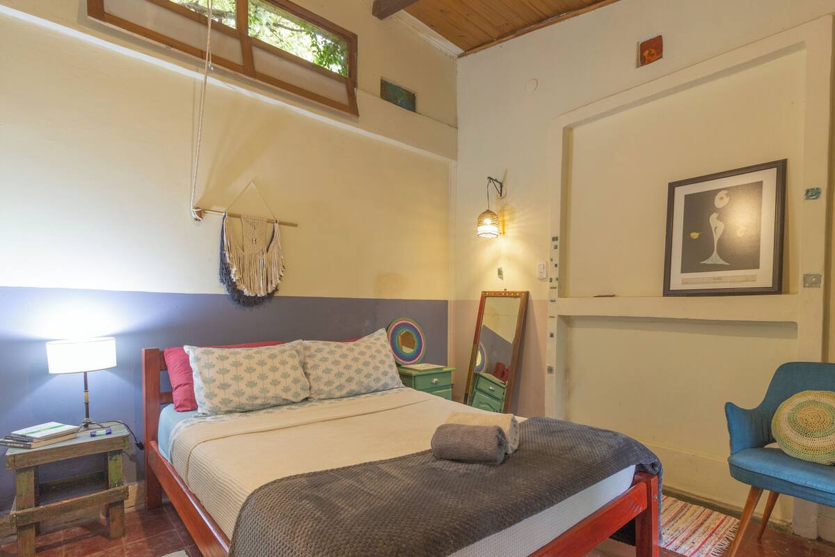 A bedroom with a bed, bedside table, lamp, dresser, chair, and artwork on the wall.