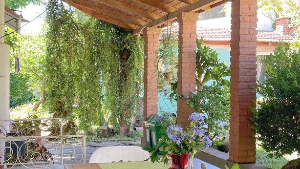 An outdoor covered patio with a wooden table, a red vase with flowers, and a teapot. - Casa del Árbol Hostel (Concepción del Uruguay)