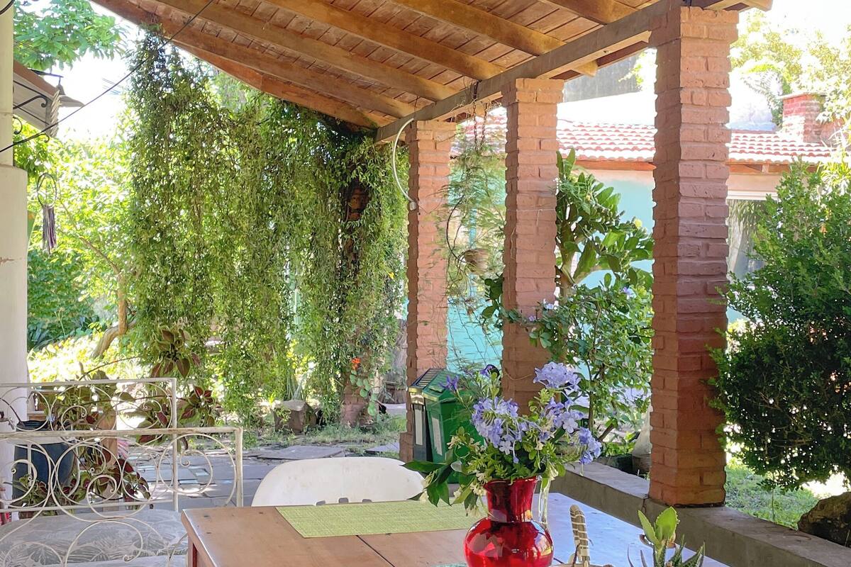 An outdoor covered patio with a wooden table, a red vase with flowers, and a teapot.