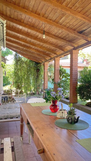 Una veranda esterna coperta con un tavolo di legno, un vaso rosso con fiori e un bollitore.
