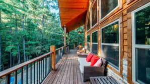 Una terraza de madera con mobiliario exterior, ventanas amplias y vistas a un bosque.