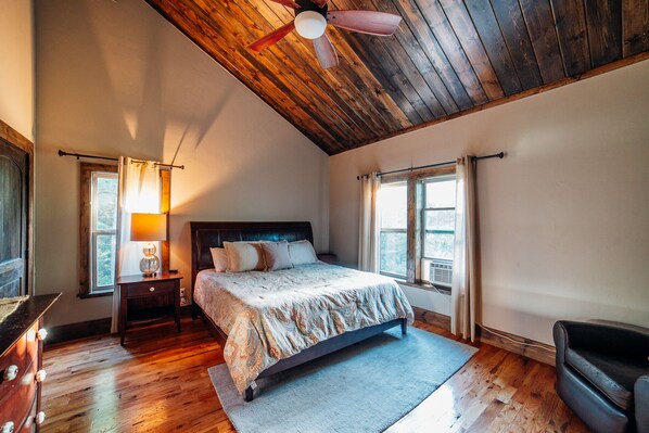 Un dormitorio con techo de madera, una cama con colcha estampada, dos mesitas de noche con lámparas y vistas al exterior por una ventana.
