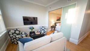 Un salon avec un canapé, un fauteuil à motifs et une petite table sur laquelle se trouve un vase avec des fleurs. Un téléviseur à écran plat est fixé au mur.