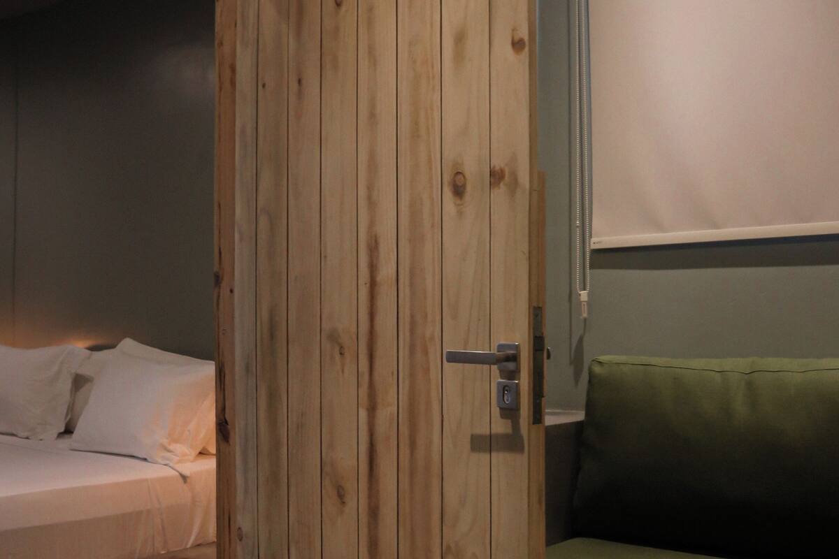 Una habitación con puerta de madera, cama con ropa de cama blanca y sofá verde.