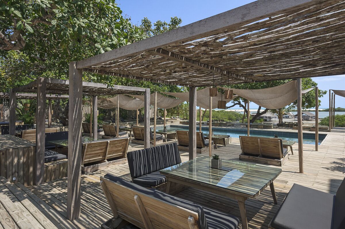 Un área de estar al aire libre con muebles de madera y una mesa de vidrio, con vistas a una piscina y una playa.