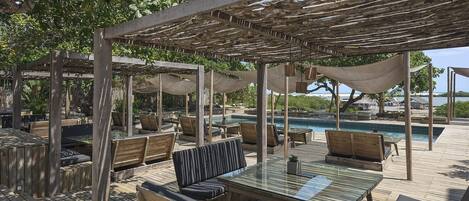 Un área de estar al aire libre con muebles de madera y una mesa de vidrio, con vistas a una piscina y una playa.