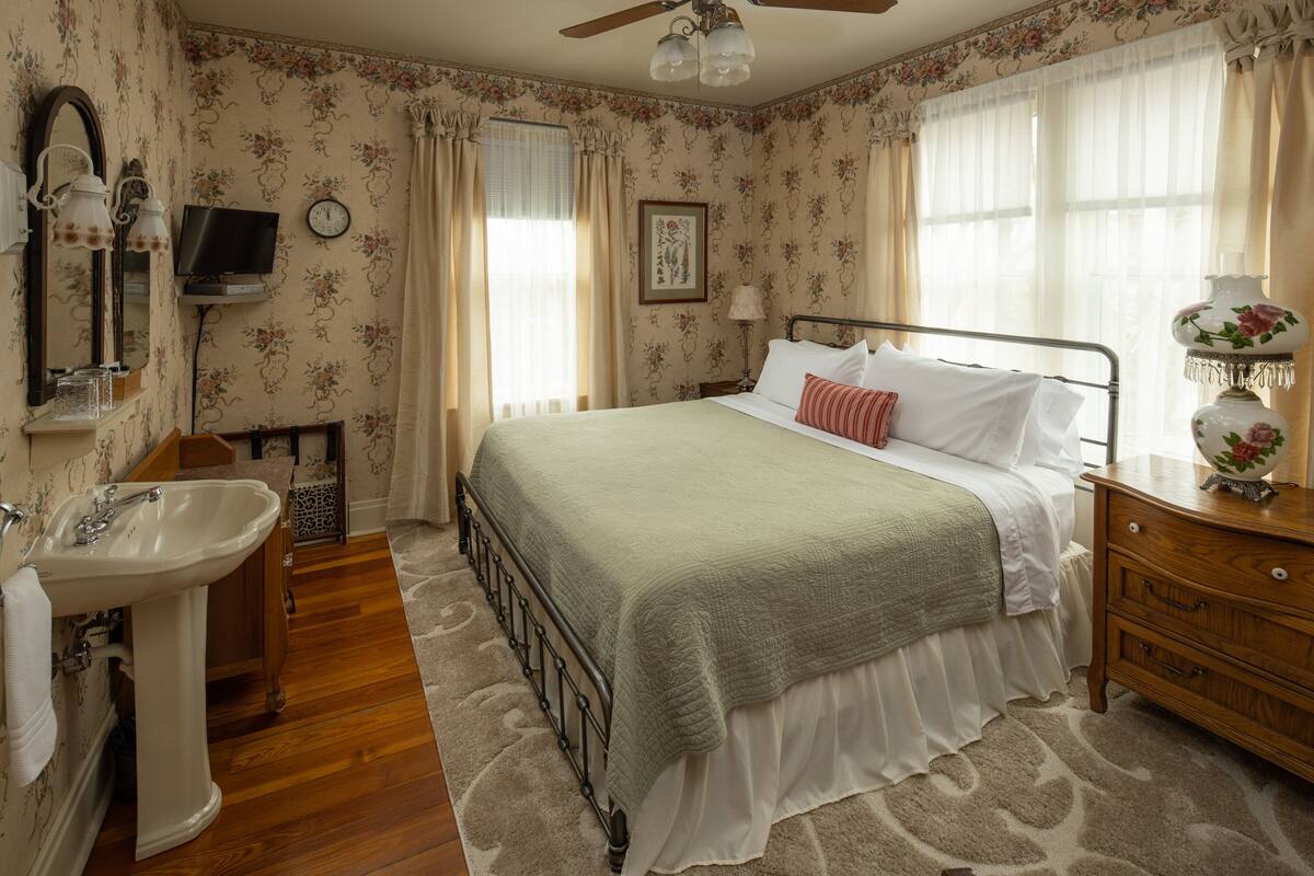 Une chambre à coucher comprenant un lit, une table de chevet, une commode, un miroir, une horloge murale et une fenêtre avec des rideaux.