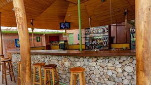 Una zona de bar con taburetes de madera, pared de piedra, barra y una pantalla de televisión.