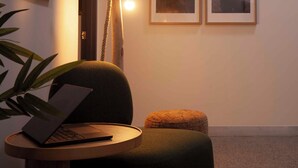 Una habitación acogedora con una laptop sobre una mesa redonda de madera, un sillón verde, un pequeño reposapiés y una lámpara fijada a la pared.