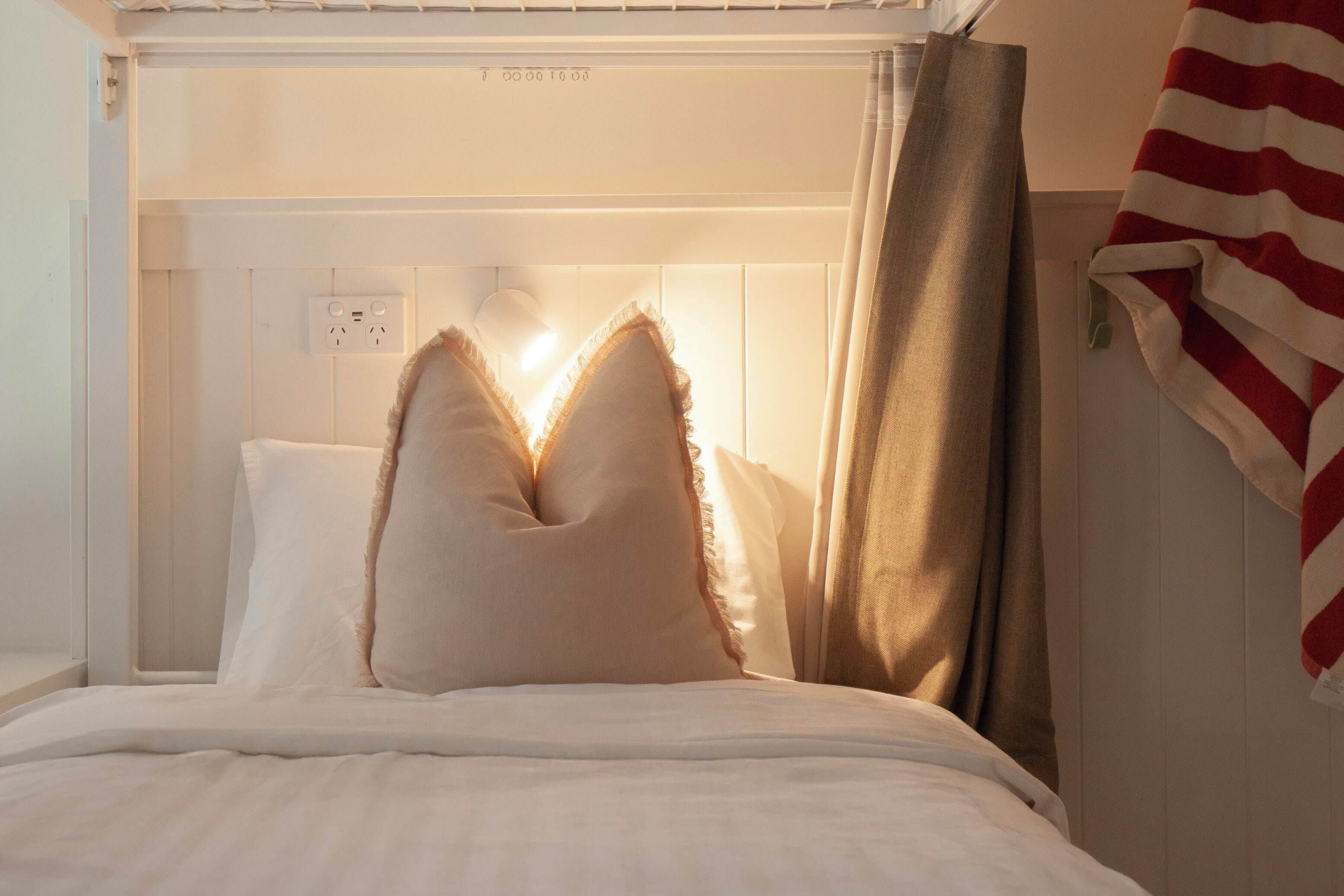 A bed with white bedding and two pillows, a window with curtains, and a power outlet on the wall.