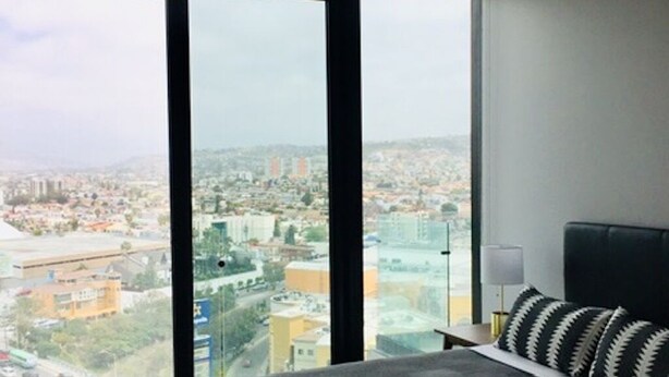 A modern bedroom with a large bed, a nightstand, and a view of a cityscape through floor-to-ceiling windows. - Loft Style Condo with Stunning View of Tijuana (Tijuana)