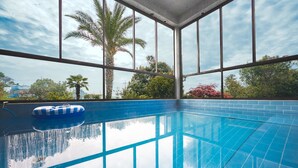 An indoor swimming pool with clear blue water, large windows, and a view of trees and a palm tree outside. - The Some Plus Onpyeong (Seogwipo)
