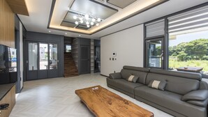 A modern living room with a wooden coffee table, grey sofa, and large windows offering a view of the outdoors. - The Some Plus Onpyeong (Seogwipo)