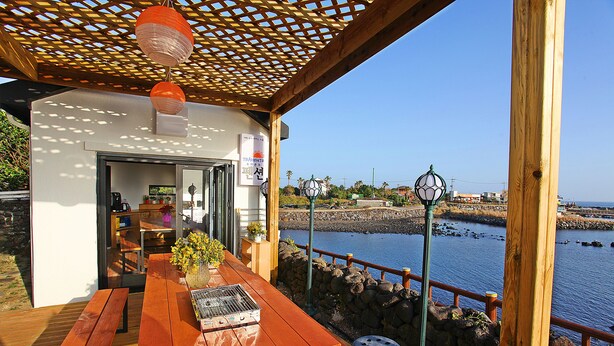 A wooden deck with a table and benches, overlooking a body of water and a stone wall. - Tramonto Pension (Seogwipo)