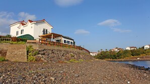 A coastal house with a white exterior and red roof, situated on a rocky shoreline with a clear blue sky. - Tramonto Pension (Seogwipo)