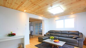 A modern living room with a wooden ceiling, a grey sofa, a coffee table with flowers, and a glass-top table with a candle. - Tramonto Pension (Seogwipo)