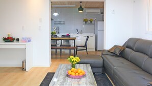 A modern living room with a grey sofa, a coffee table with a vase of flowers, and a view into the kitchen area. - Tramonto Pension (Seogwipo)
