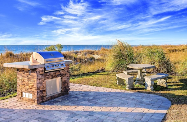 Un espace barbecue extérieur avec un gril en pierre, une table et deux chaises, entouré de pelouse et offrant une vue sur l’océan.