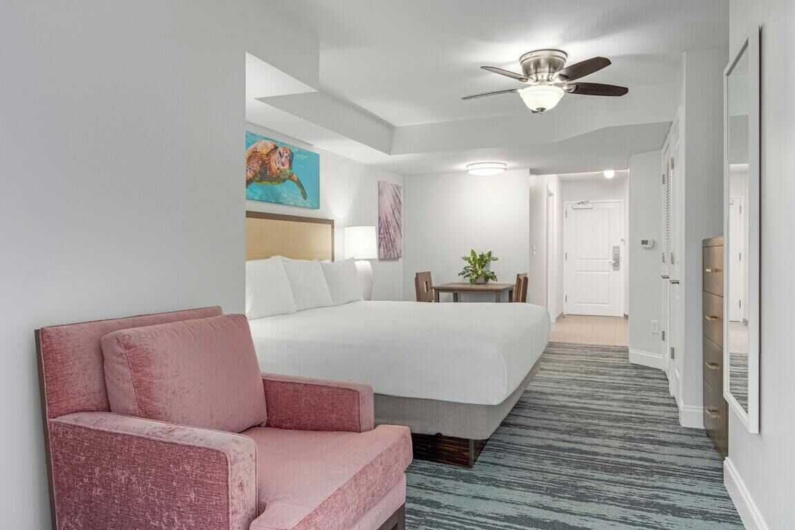 Quarto de hotel com cama, uma cadeira rosa, ventilador de teto, quadro e uma pequena mesa com uma planta.
