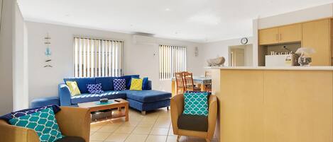 A modern living room with a blue sofa, two armchairs, a wooden coffee table, and a kitchen area with a counter and cabinets.