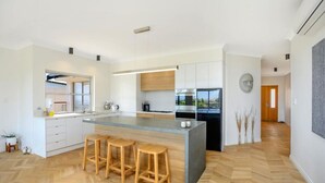 Une cuisine moderne dotée d’un îlot central, de tabourets en bois, de meubles blancs et offrant une vue sur l’extérieur.