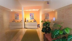 Et hotellets receptionområde med et grønt tæppe, der fører til en receptionsdisk, to receptionist og en stor stueplante.