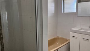 Kylpyhuoneessa on valkoiset laatat, lasinen suihkuseinä, beigenvärinen kylpyamme ja valkoinen pesuallas, jonka yläpuolella on peili.
