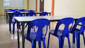 Une salle de classe avec des chaises en plastique bleues, un tableau blanc et un ventilateur au plafond.