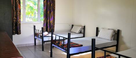 A dormitory room with three bunk beds, a wooden desk, a window with curtains, and an air conditioning unit.
