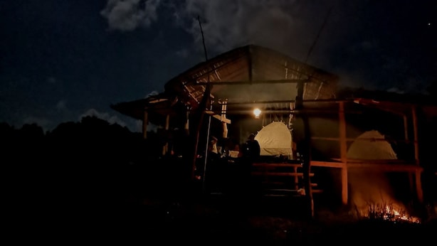 Une cabane rustique avec un toit de chaume, un petit feu qui crépite à l’intérieur, et une personne debout près de l’entrée.