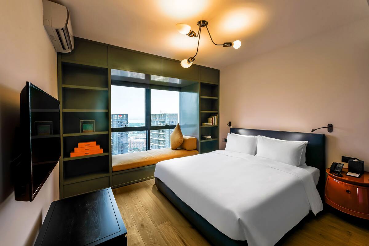 Habitación de hotel con una cama grande, estantes integrados, un ventanal con vistas a la ciudad y una mesita de noche con teléfono y lámpara.