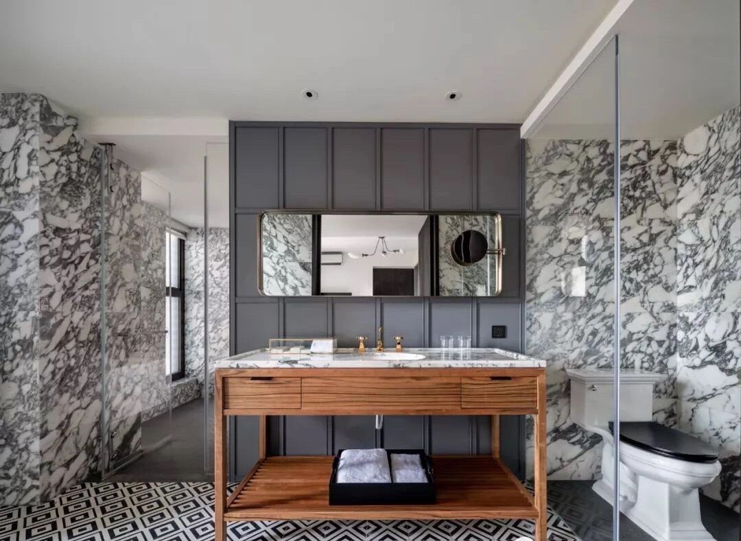 A modern bathroom with a marble wall and floor, a wooden vanity with a mirror, and a toilet.