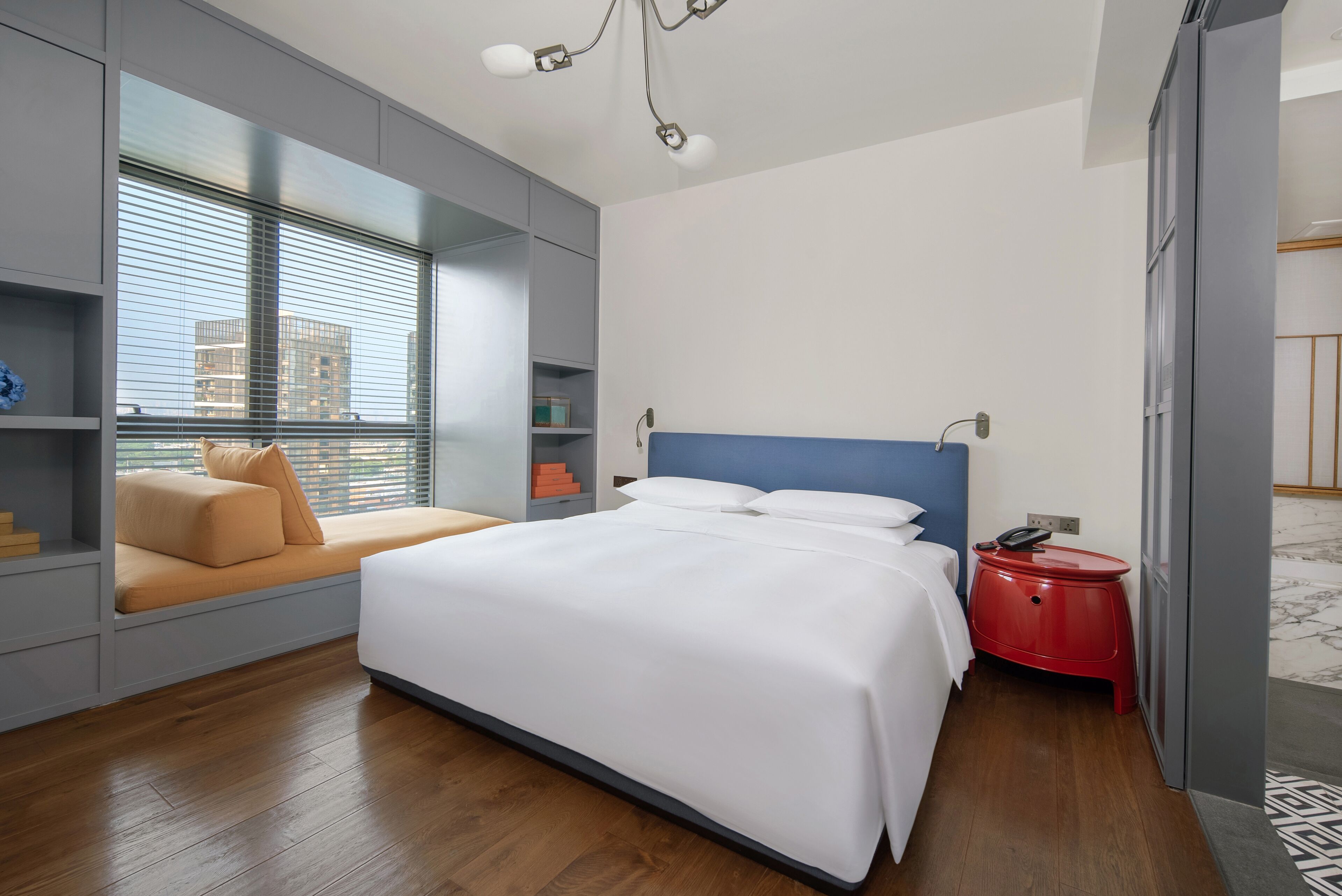 A modern hotel room with a large bed, a sofa bed, a red side table, and a view of the city through floor-to-ceiling windows.