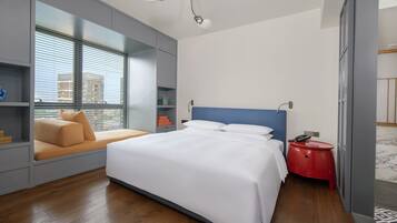 A modern hotel room with a large bed, a sofa bed, a red side table, and a view of the city through floor-to-ceiling windows.