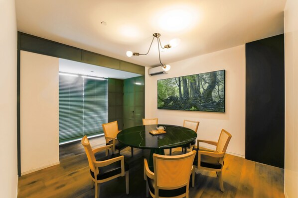 Uma sala de conferência com uma mesa redonda, seis cadeiras, um projetor e um quadro com uma paisagem de floresta na parede.