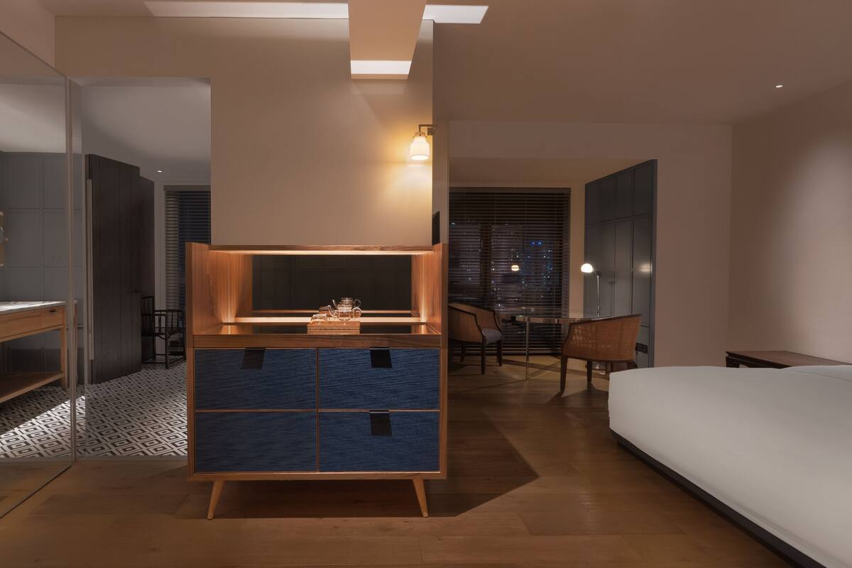 Una habitación de hotel moderna con una cama grande, un mueble de madera con cajones, un escritorio con sillas y un espejo grande.