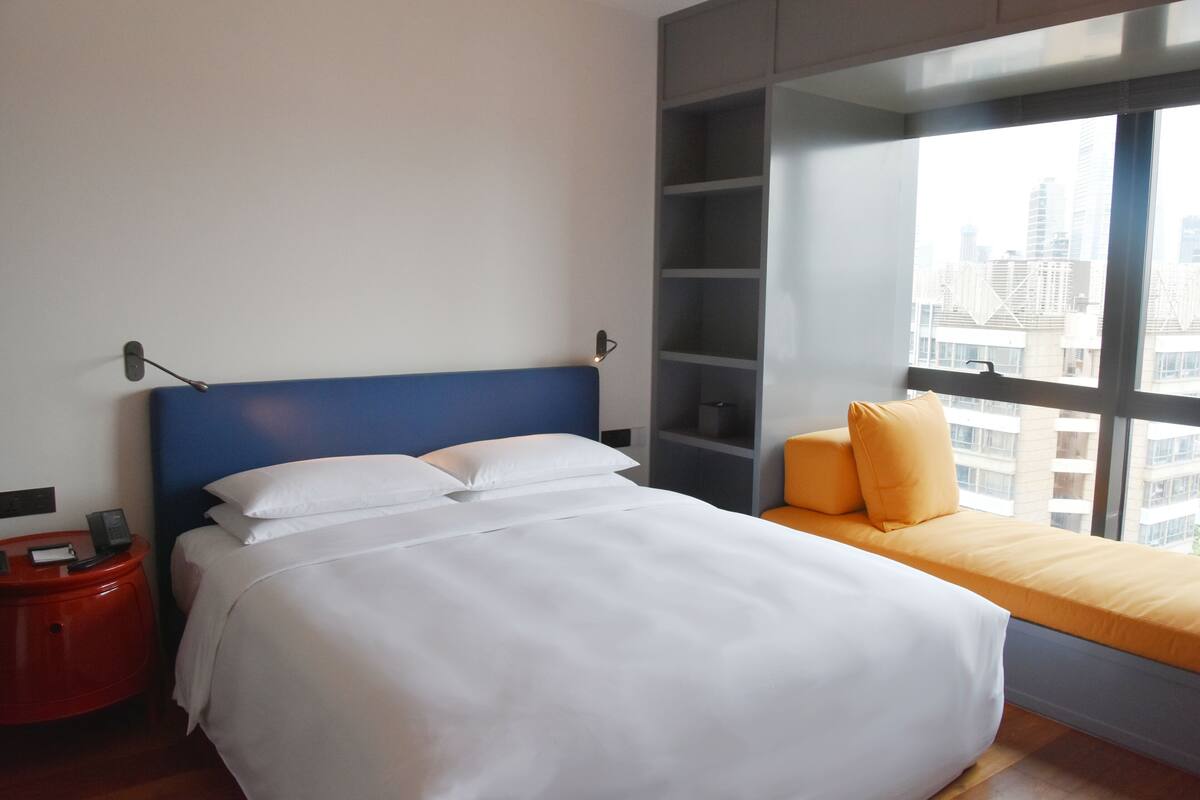 Habitación de hotel con cama, una mesita de noche roja, ventana con vista a la ciudad y cabecero azul.
