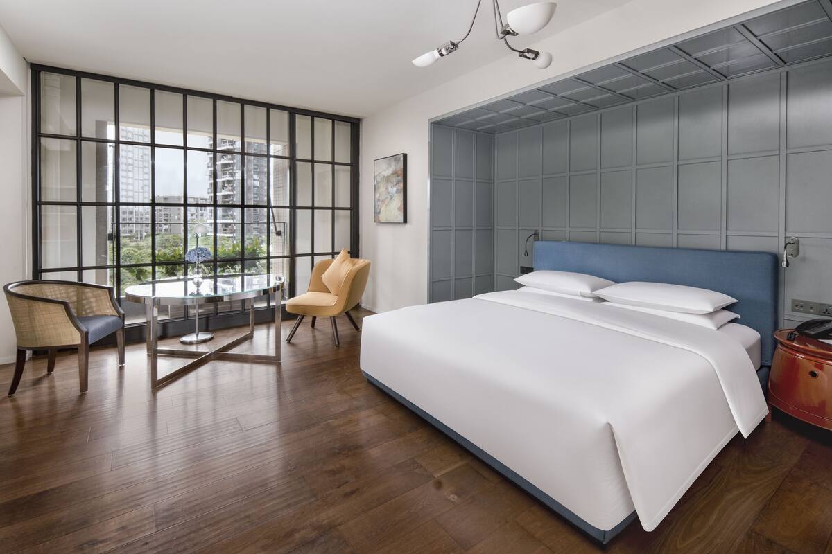 Habitación de hotel moderna con una cama grande, una zona de estar con mesa y sillas, y vistas de la ciudad a través de amplias ventanas.