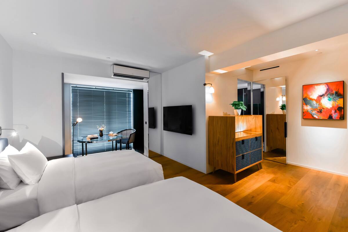 Habitación de hotel moderna con cama, escritorio y televisor. Hay una ventana con persianas, una planta y un cuadro en la pared.
