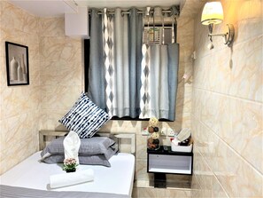 A hotel room with a bed, a bedside table, a nightstand, a lamp, a mirror, a window with curtains, and a wall-mounted air conditioning unit. - Super Guest House (Kowloon)