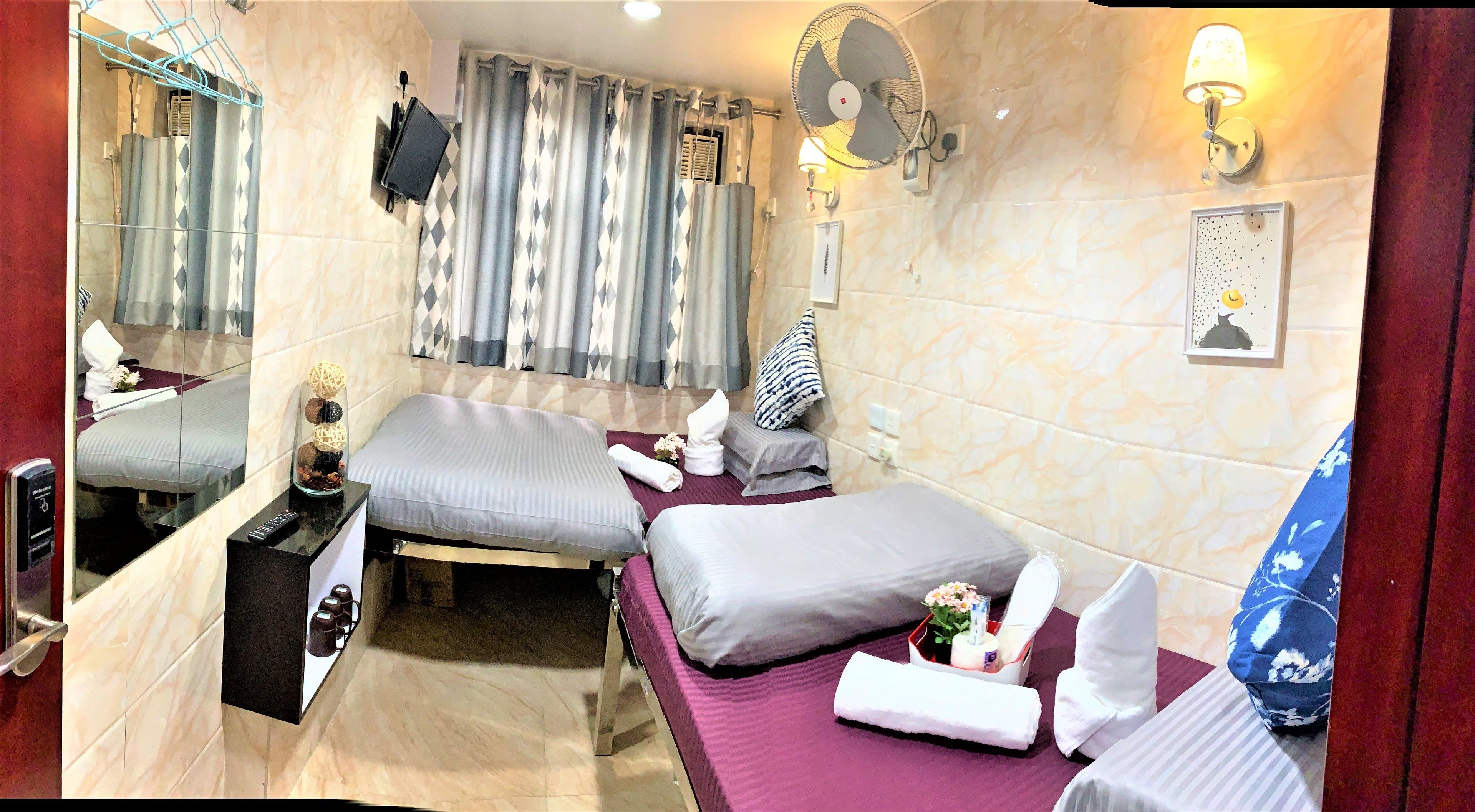 Habitación de hotel con dos camas, baño con ducha, espejo, ventilador e iluminación en la pared.