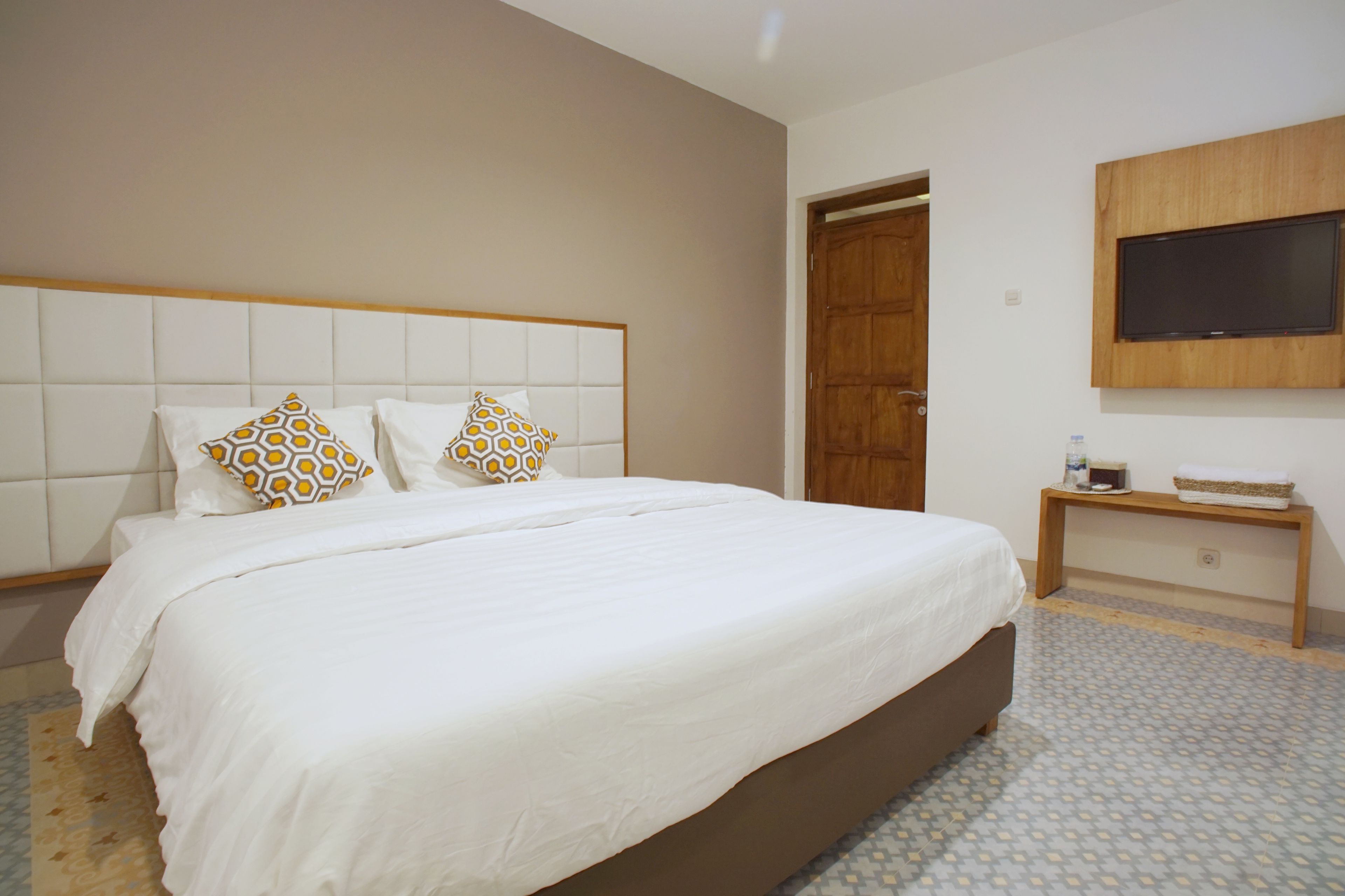 A bedroom with a large bed, two patterned pillows, a wooden bedside table with a bottle and tissue box, and a TV mounted on the wall.